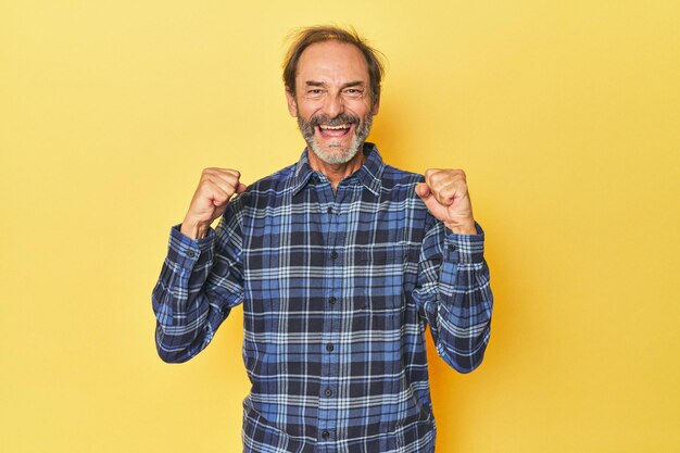 Hombre caucásico de mediana edad en un estudio amarillo animando el concepto de victoria despreocupado y emocionado