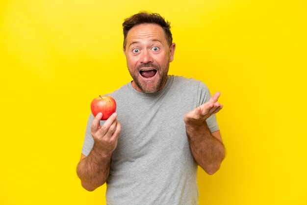 Hombre caucásico de mediana edad con una manzana aislada de fondo amarillo con una expresión facial sorprendida