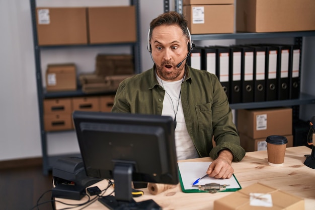 Hombre caucásico de mediana edad que trabaja en el comercio electrónico de pequeñas empresas con auriculares asustados y asombrados con la boca abierta por la cara sorpresa de incredulidad