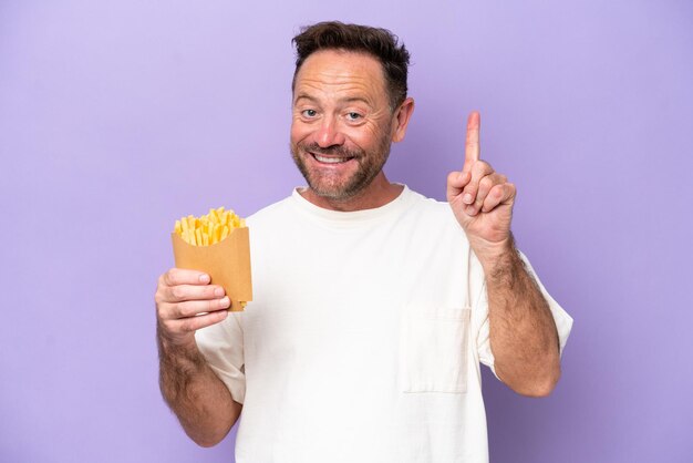 Hombre caucásico de mediana edad sosteniendo papas fritas aisladas en un fondo morado mostrando y levantando un dedo en señal de lo mejor