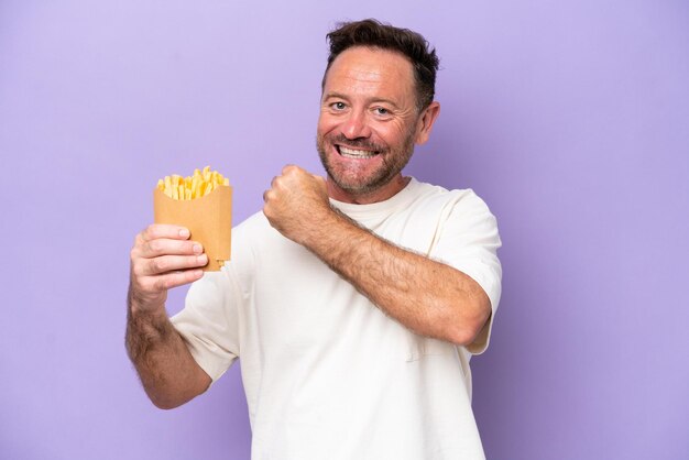 Hombre caucásico de mediana edad sosteniendo papas fritas aisladas en un fondo púrpura celebrando una victoria