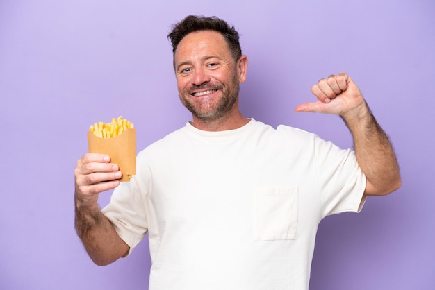 Hombre caucásico de mediana edad sosteniendo papas fritas aisladas en un fondo púrpura orgulloso y satisfecho de sí mismo