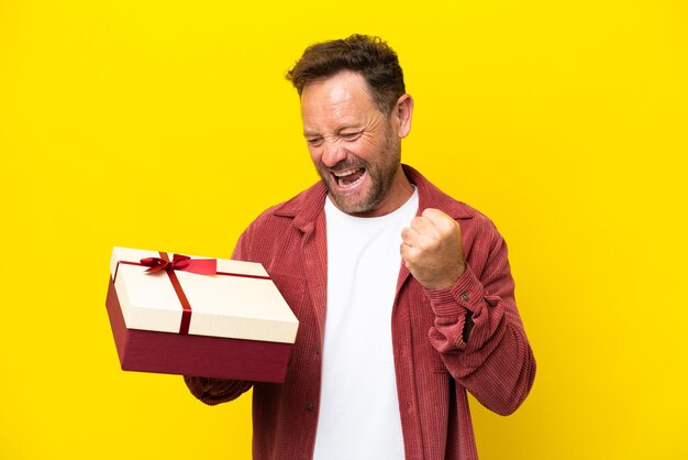 Hombre caucásico de mediana edad sosteniendo un regalo aislado de fondo amarillo celebrando una victoria