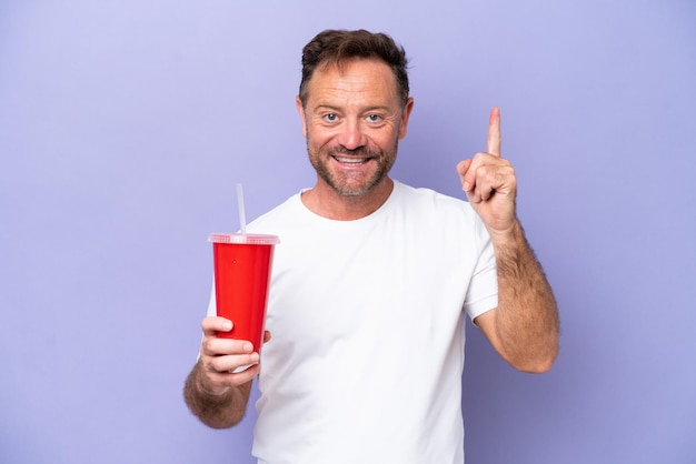 Hombre caucásico de mediana edad sosteniendo soda aislada en un fondo morado mostrando y levantando un dedo en señal de lo mejor