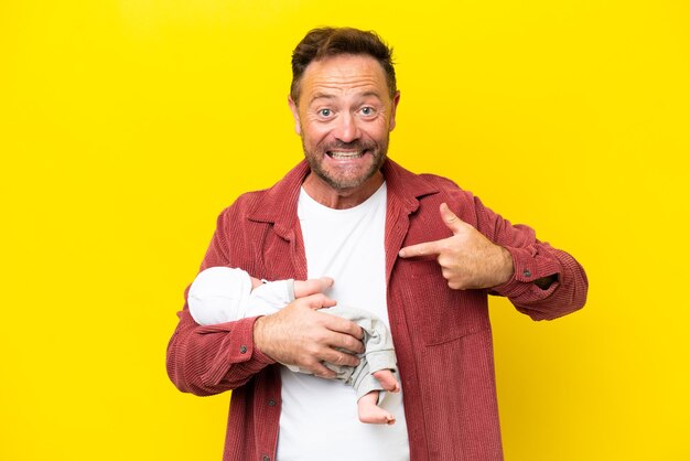 Hombre caucásico de mediana edad sosteniendo a su hijo recién nacido aislado de fondo amarillo con expresión facial sorpresa