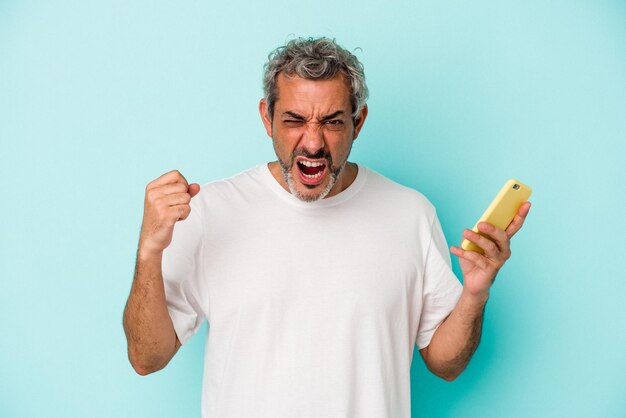 Hombre caucásico de mediana edad sosteniendo un teléfono móvil aislado de fondo azul gritando muy enojado y agresivo.