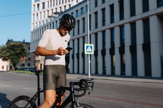 Hombre caucásico musculoso con casco de ropa deportiva y gafas de pie en la calle de la ciudad con bicicleta negra y usando un teléfono inteligente moderno Descansando después del entrenamiento