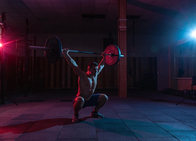 Hombre caucásico musculoso con cuerpo musculoso haciendo prensa de pesas en un gimnasio sucio oscuro con luz de neón roja azul