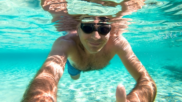 Hombre caucásico nadando bajo el agua en gafas de natación agua azul transparente