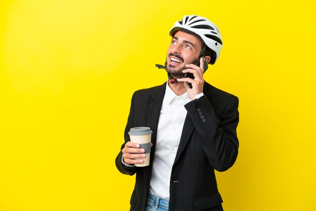 Hombre caucásico de negocios con un casco de bicicleta aislado en un fondo amarillo sosteniendo café para llevar y un móvil