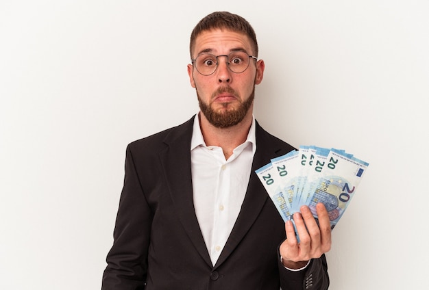 Foto hombre caucásico de negocios joven con billetes aislados sobre fondo blanco se encoge de hombros y abre los ojos confundidos.