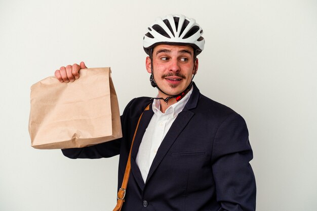 Hombre caucásico de negocios joven con casco de bicicleta y sosteniendo comida de manera aislada sobre fondo blanco se ve a un lado sonriente, alegre y agradable.