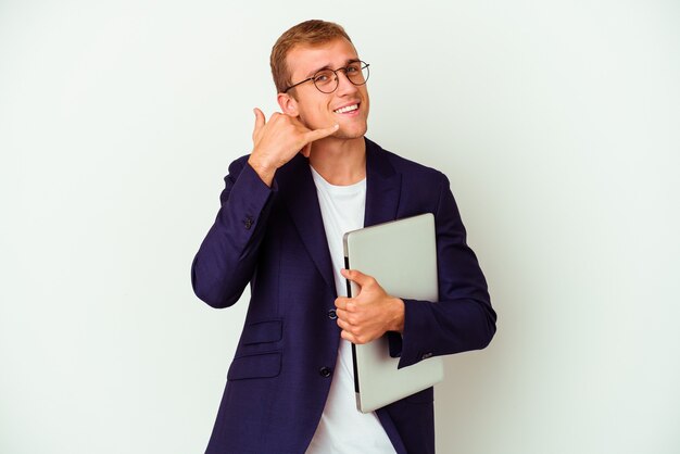 Hombre caucásico de negocios joven que sostiene una computadora portátil aislada en blanco que muestra un gesto de la llamada del teléfono móvil con los dedos.