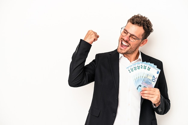 Hombre caucásico de negocios joven sosteniendo billetes aislados sobre fondo blanco levantando el puño después de una victoria, concepto ganador.