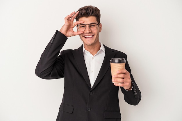 Hombre caucásico de negocios joven sosteniendo café para llevar aislado sobre fondo blanco emocionado manteniendo el gesto de ok en el ojo.