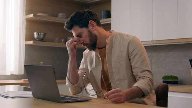 Hombre caucásico ocupado y independiente que trabaja lejos de casa trabajando con una computadora portátil en la cocina agotado