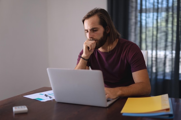 Foto hombre caucásico pensativo sentado en la mesa trabajando desde casa y usando una computadora portátil