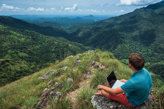 Hombre caucásico con portátil sentado en el borde de la montaña ella con impresionantes vistas del valle en Sri Lanka