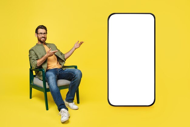 Hombre caucásico positivo sentado casualmente en una silla con un enorme teléfono celular apuntando a una maqueta de pantalla blanca vacía
