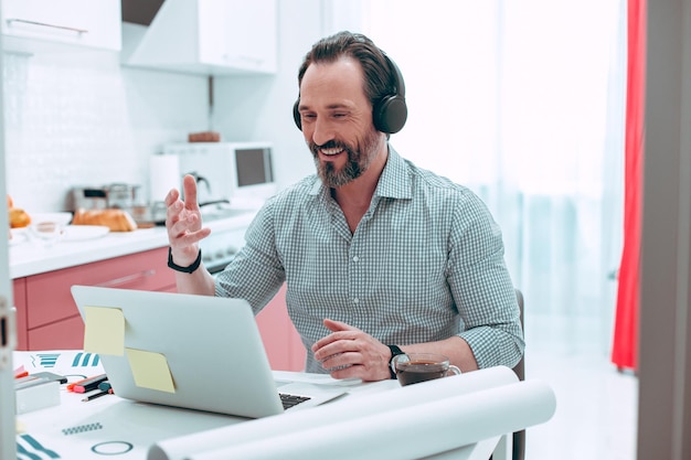 Hombre caucásico positivo sentado en la mesa de la cocina con una computadora portátil y gesticulando durante la videollamada Gráficos gráficos a su lado