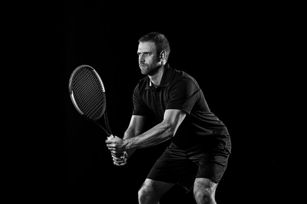 El hombre caucásico que juega al tenis aislado sobre fondo negro. Disparo monocromo de estudio de joven jugador en forma en el estudio en movimiento o movimiento durante el juego deportivo.