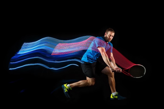 Foto el hombre caucásico que juega al tenis aislado sobre fondo negro. foto de estudio de un joven jugador en forma en el estudio en movimiento o movimiento durante el juego deportivo con rastro de luz led