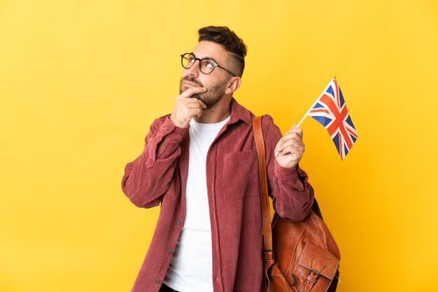 Hombre caucásico que sostiene una bandera del Reino Unido aislada en la pared amarilla que tiene dudas