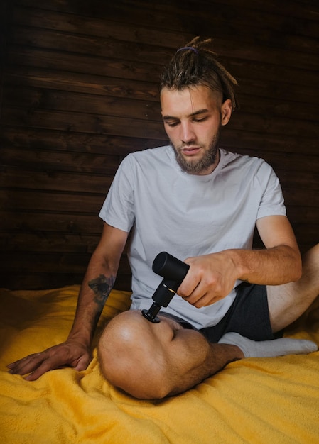 Hombre caucásico con rastas se da un masaje con pistola de percusión en casa sentado en la cama