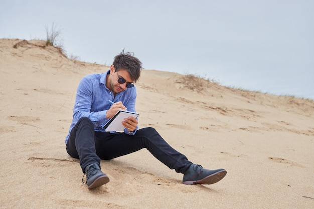 Hombre caucásico sentado en la arena de la playa escribiendo poesía concentrada o una carta. Lleva gafas de sol, camisa azul, pantalón negro y zapatos negros.