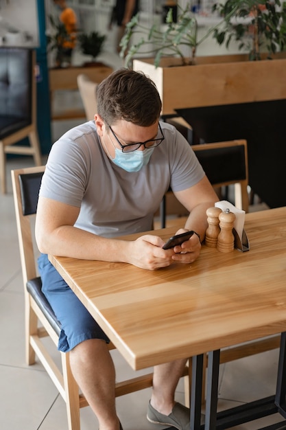 Hombre caucásico sentado en el restaurante y usando el teléfono en la mascarilla médica. Nuevo concepto normal