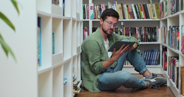 El hombre caucásico sentado en el suelo cerca de los estantes con un libro en la biblioteca usa una tableta digital y escribe notas para la tarea de clase