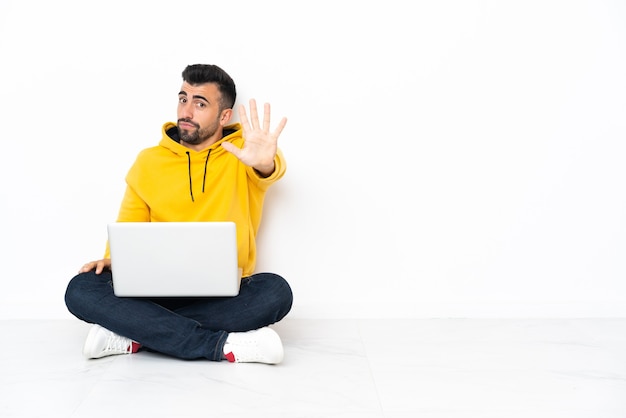 Hombre caucásico sentado en el suelo con su computadora portátil contando cinco con los dedos