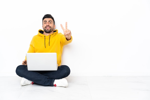 Hombre caucásico sentado en el suelo con su computadora portátil sonriendo y mostrando el signo de la victoria