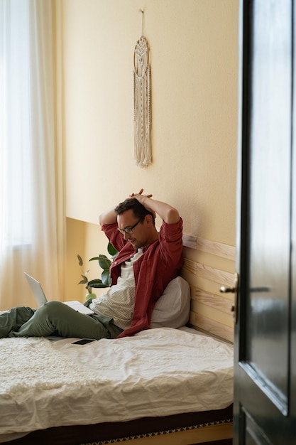 Un hombre caucásico serio y relajado mira atentamente la pantalla de un portátil sentado en la cama con las manos detrás de la cabeza