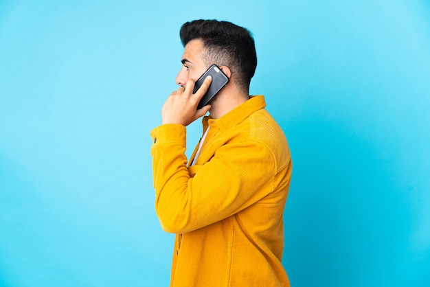 Hombre caucásico sobre pared azul aislada manteniendo una conversación con el teléfono móvil con alguien