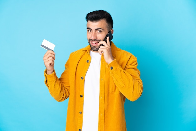 Hombre caucásico sobre pared azul aislada manteniendo una conversación con el teléfono móvil y sosteniendo una tarjeta de crédito