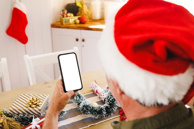 Hombre caucásico con sombrero de Papá Noel, sentado a la mesa en la cocina, usando un teléfono inteligente con espacio de copia