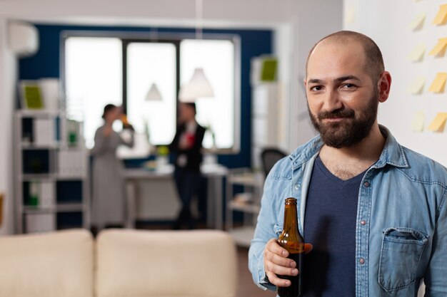 Hombre caucásico sonriendo y sosteniendo una botella de cerveza después del trabajo en la fiesta de la oficina. Los colegas se reúnen para disfrutar de actividades divertidas mientras juegan juegos, comen, beben alcohol. Bebidas de celebración