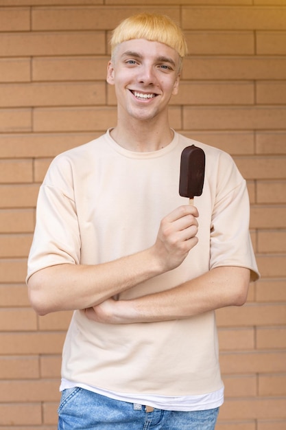 Un hombre caucásico sonriente parado en una camiseta beige y sosteniendo un helado de chocolate en un palo