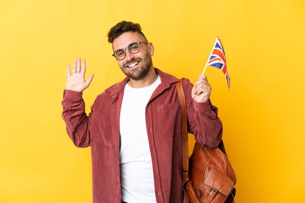 Hombre caucásico sosteniendo una bandera del Reino Unido aislado sobre fondo amarillo saludando con la mano con expresión feliz