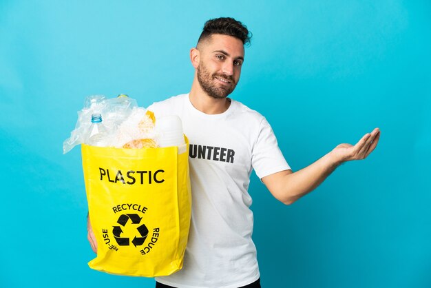Hombre caucásico sosteniendo una bolsa llena de botellas de plástico para reciclar aislado sobre fondo azul extendiendo las manos hacia el lado para invitar a venir