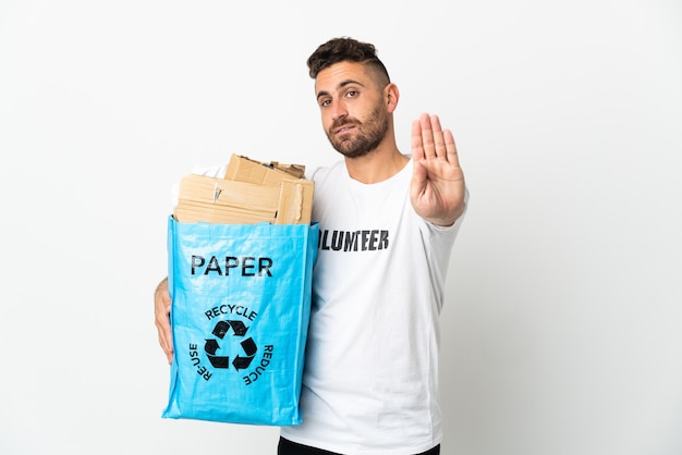 Hombre caucásico sosteniendo una bolsa de reciclaje llena de papel para reciclar aislado sobre fondo blanco haciendo gesto de parada