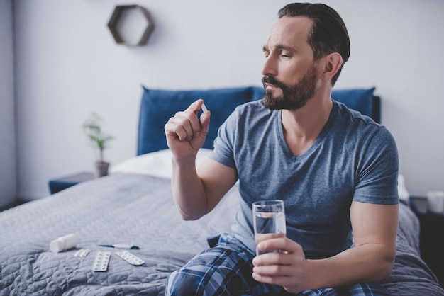 Hombre caucásico sosteniendo una pastilla y un vaso de agua sentado en la cama