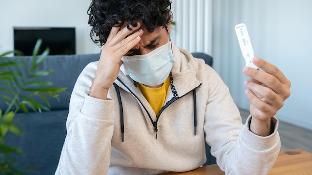 Un hombre caucásico triste hace un dispositivo de prueba rápido en el hogar para el virus Covid19 con un resultado positivo y sostiene su cabeza en la mesa de madera Casete de antígeno de pcr para el nuevo virus variante Omicron