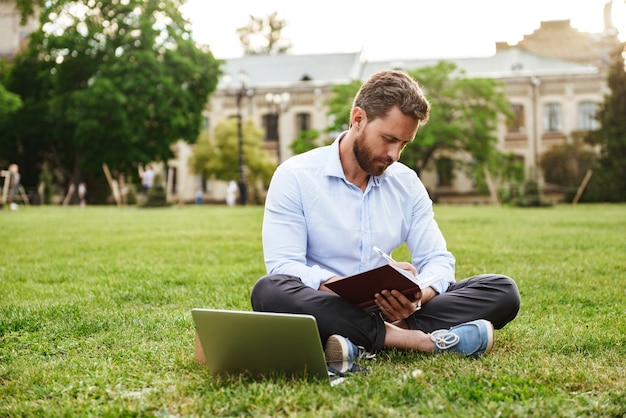 Hombre caucásico vestido con camisa blanca, sentado en el césped en el parque con las piernas cruzadas y anotando notas en el cuaderno mientras trabaja en la computadora portátil