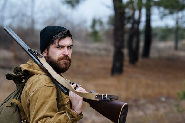 Foto un hombre cazador con barba y bigote en un sombrero negro y una gran mochila de senderismo sostiene una pistola en su hombro en el bosque