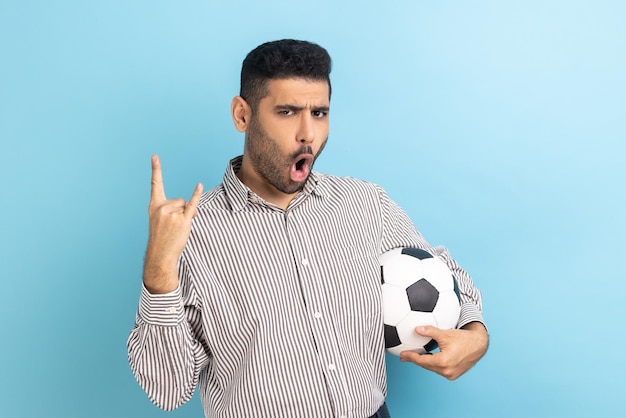 Hombre celebrando la victoria del equipo de fútbol favorito sosteniendo un balón de fútbol en las manos mostrando un gesto de rock