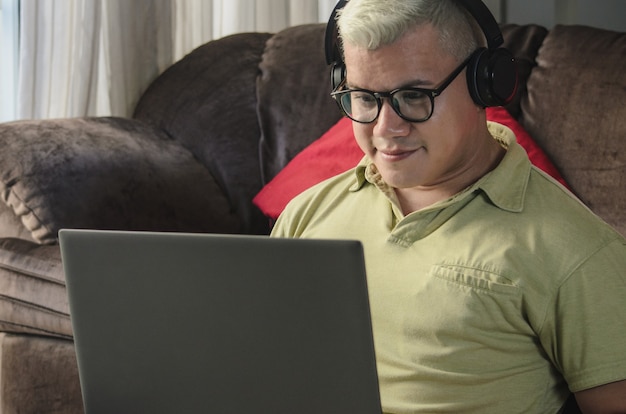 Hombre de cerca con gafas sentado en el piso de la sala junto al sofá, comprobando el portátil con auriculares