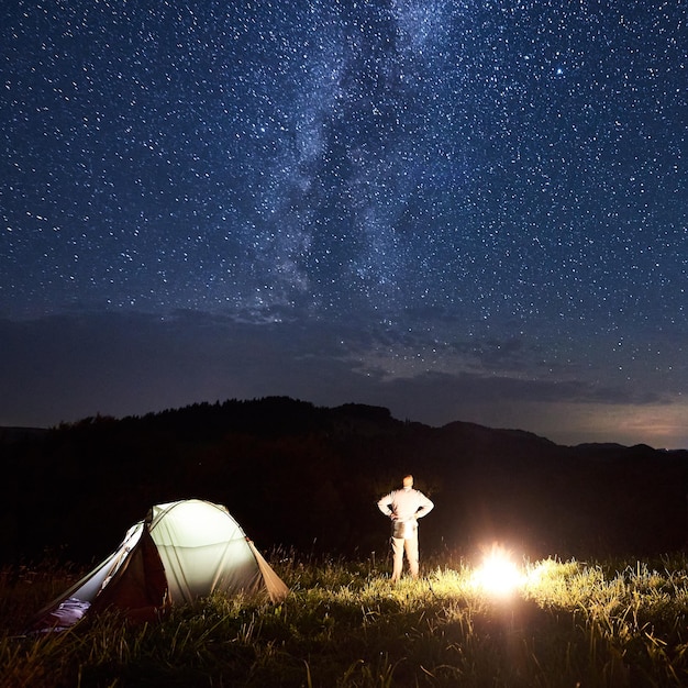 El hombre cerca de la tienda y la hoguera se encuentra con la Vía Láctea en el cielo estrellado contra el fondo de las montañas