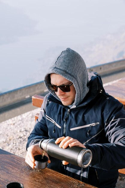 Foto un hombre con una chaqueta con una capucha tirada vierte café de un termo en una taza mientras está sentado en una mesa